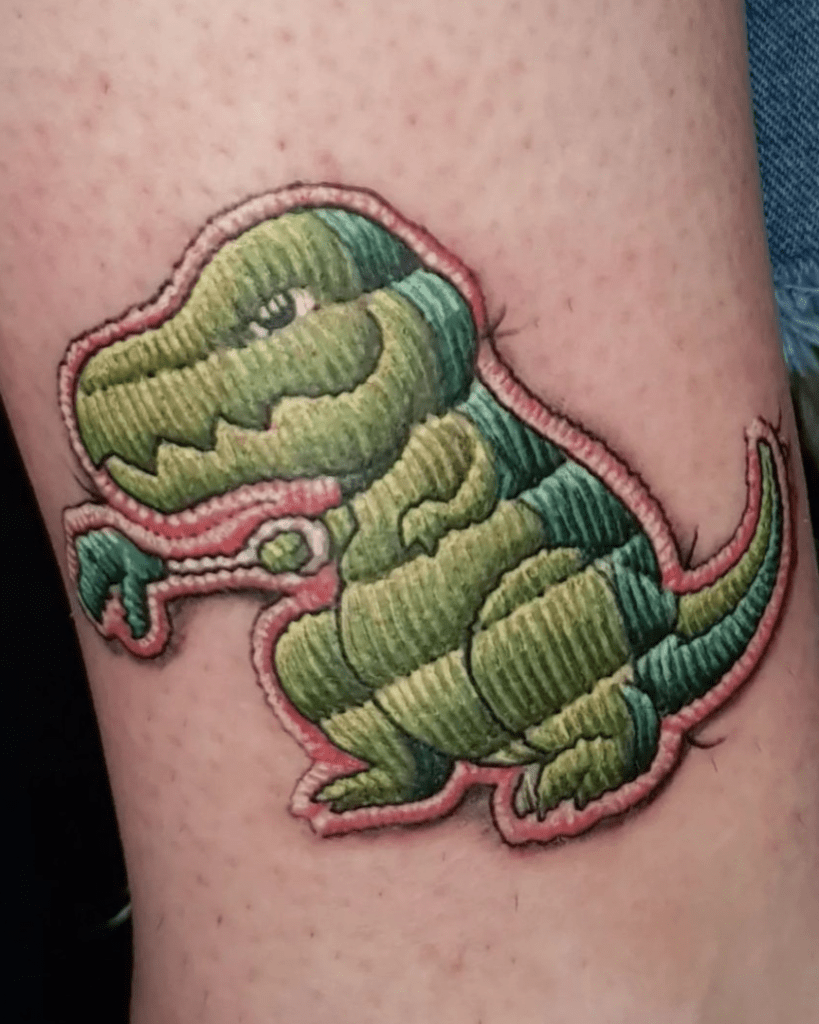 Artista: Piero Tat-Twin. Pro Team Artist Water Law Tattoo. Tatuaggio realistico a colori di una toppa ricamata di colore verde raffigurante un tirannosauro.